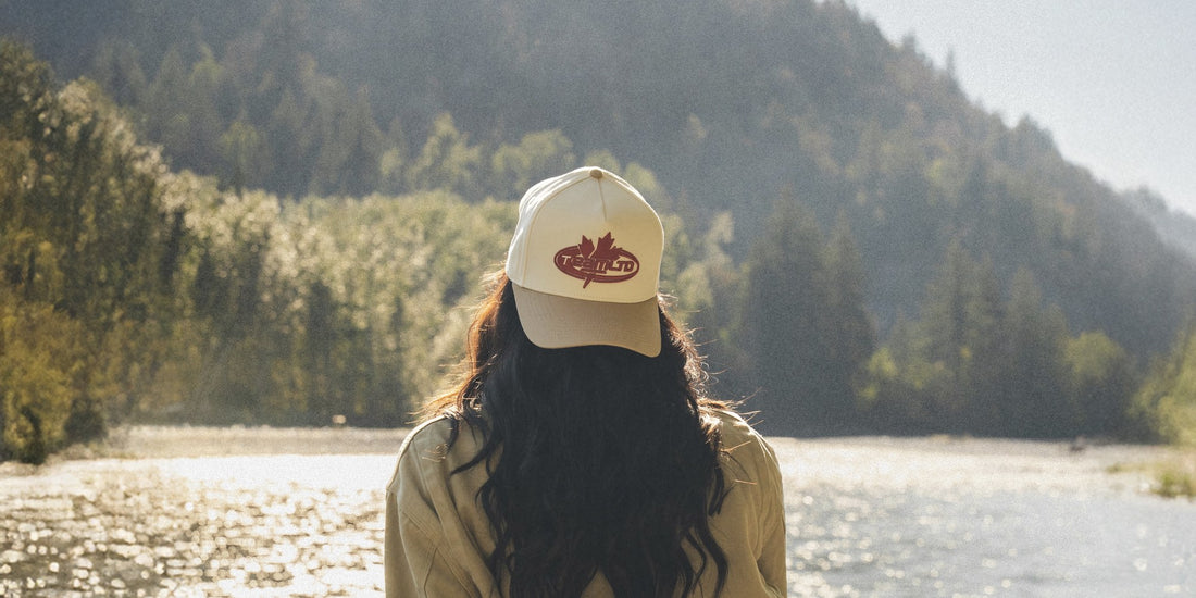 A woman in the cream and sand TEAMLTD Shield Snapback sits by a river below a forested mountain